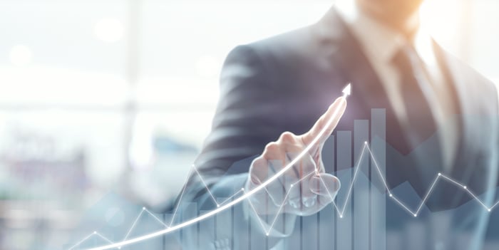 A businessman draws an upward arrow over a stock chart displayed on a transparent touchscreen.