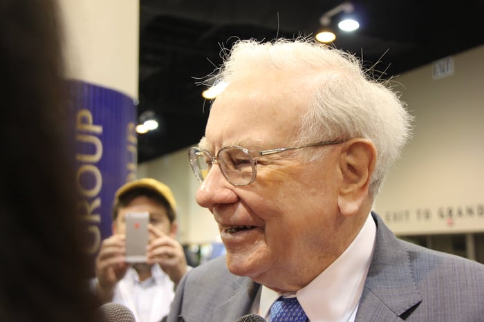Warren Buffett smiling with a person in the background taking his photo using a cell phone.